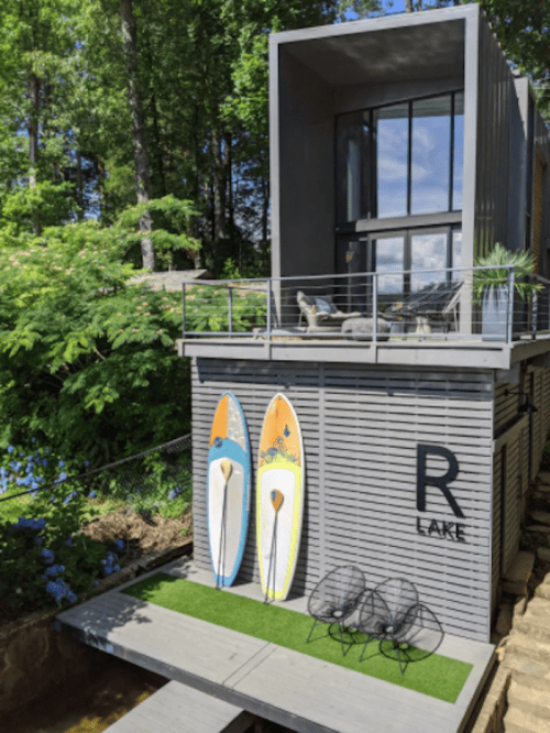 Modern lakeside house with large windows, two paddleboards, and outdoor seating surrounded by greenery.