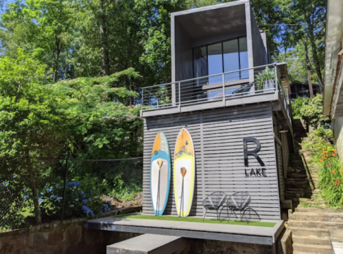 Modern lakeside house with large windows, two colorful paddleboards, and a small patio surrounded by greenery.