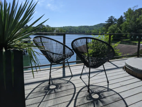 Two black chairs on a deck overlooking a serene lake, surrounded by greenery and a clear blue sky.