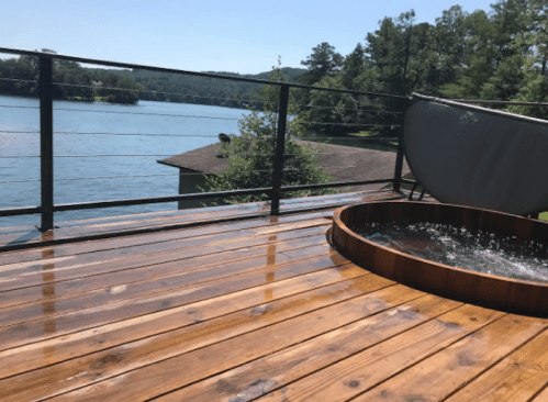 A wooden deck overlooking a lake, featuring a round hot tub and lush greenery in the background under a clear blue sky.