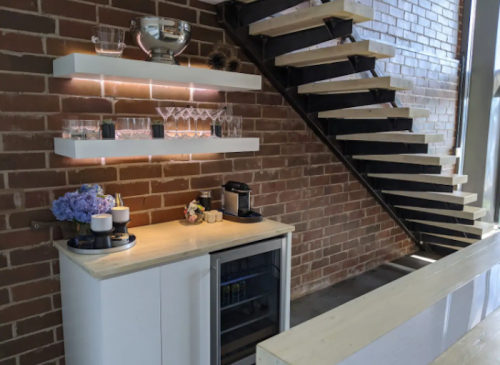 A modern kitchen corner with a mini fridge, glassware, and flowers, beside a stylish staircase against a brick wall.