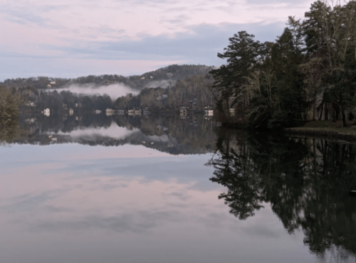 Serene lake scene at dawn, with misty hills and reflections of trees and houses on calm water.