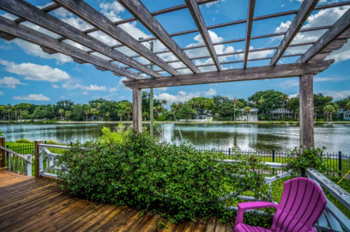 A scenic view of a lake framed by a wooden pergola, with lush greenery and a pink chair on a wooden deck.