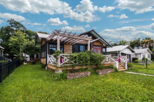 A charming wooden house with a porch, surrounded by greenery and under a bright, cloudy sky.