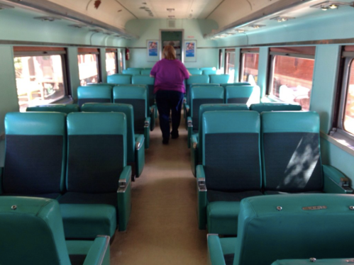 A person walks down the aisle of a vintage train car with teal seats and large windows.