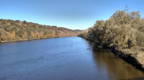 A serene river flows through a landscape of autumn trees under a clear blue sky.