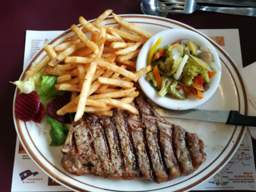 A plate with grilled steak, French fries, and a small bowl of mixed pickled vegetables, garnished with lettuce and beets.