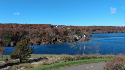 Scenic view of a lake surrounded by autumn foliage and a clear blue sky. A path runs along the water's edge.