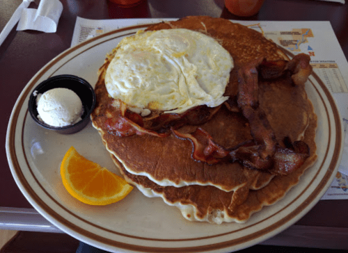 A plate of pancakes topped with a fried egg, bacon, a slice of orange, and a small cup of butter.