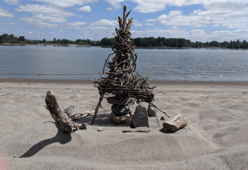 A small sculpture made of sticks and stones on a sandy beach, with a river and trees in the background under a blue sky.
