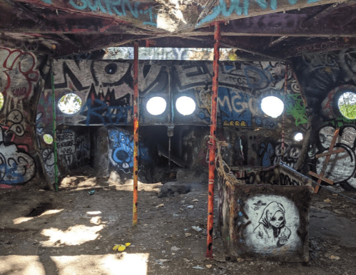 Interior of a graffiti-covered abandoned structure with circular windows and a dark, shadowy area in the corner.