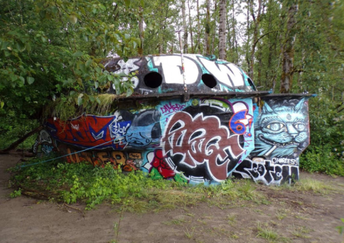 A graffiti-covered abandoned structure surrounded by trees and greenery, with a sandy ground in the foreground.