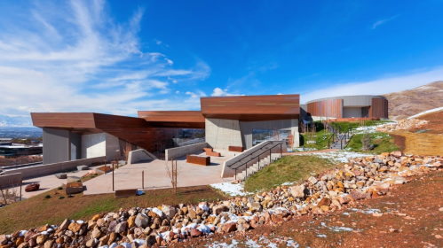 Modern architectural building with wooden and concrete elements, surrounded by rocky terrain and snow, under a blue sky.