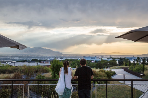 A couple stands on a balcony, gazing at a scenic view of mountains and a sunset with rays of light breaking through clouds.