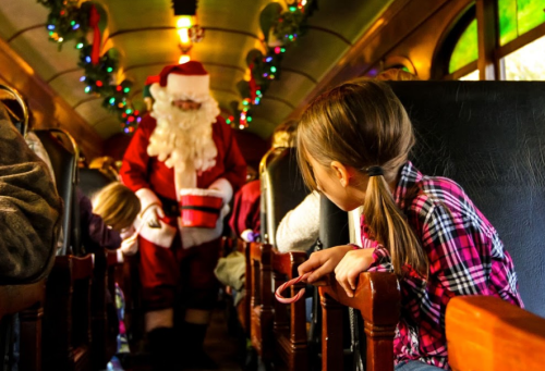 Santa Claus interacts with children on a festive train, decorated with holiday lights and greenery.