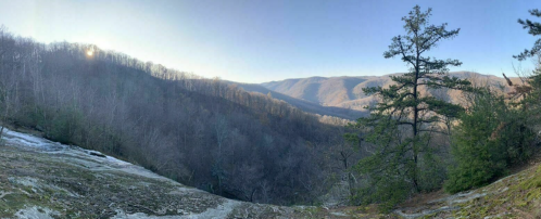 Panoramic view of a mountainous landscape with trees, a waterfall, and a clear blue sky at sunset.