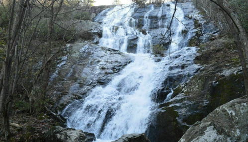 A cascading waterfall flows over rocky terrain, surrounded by trees and natural scenery.