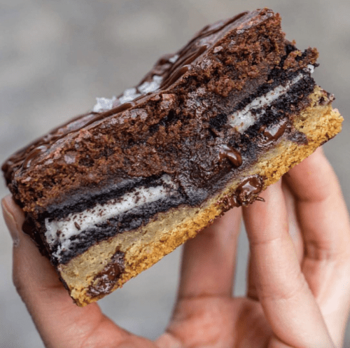 A hand holding a layered dessert with chocolate brownie, Oreo, and cookie dough layers, topped with chocolate drizzle.