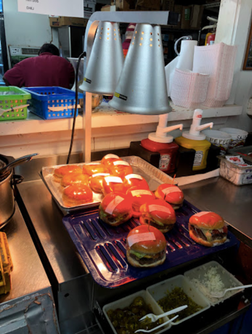 A countertop with burgers under heat lamps, condiments nearby, and a kitchen area in the background.