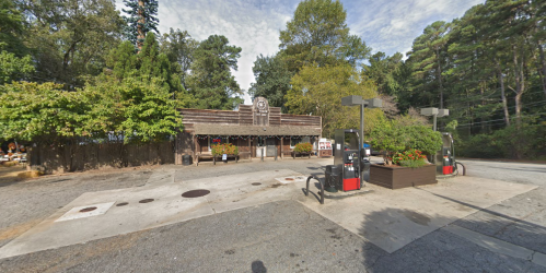 A rustic gas station with a wooden building, surrounded by trees and flower planters, on a quiet road.