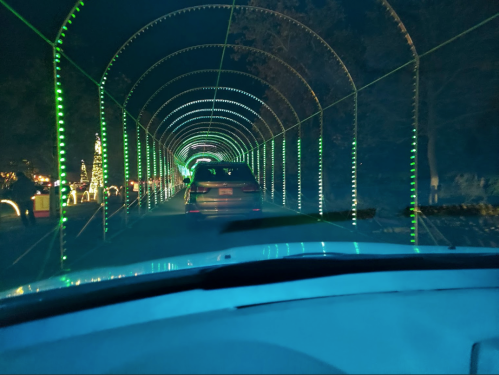 A car drives through a tunnel of green lights at night, surrounded by festive decorations.