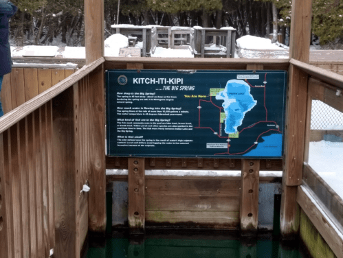 Sign at Kitch-iti-kipi, the Big Spring, with a map and information about the spring's features and history. Snowy surroundings.