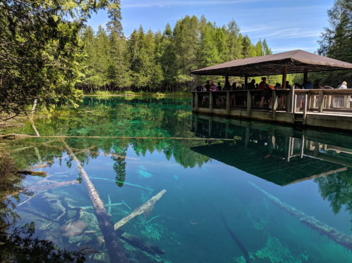 A serene lake with clear blue water, surrounded by trees, featuring a wooden observation deck with visitors.