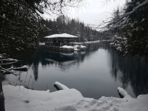 A serene winter scene featuring a snowy landscape, a calm lake, and a small gazebo on the water's edge.