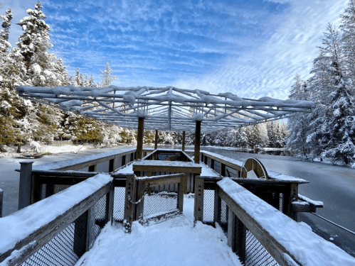Snow-covered wooden structure with a canopy, surrounded by trees and a frozen lake under a blue sky.