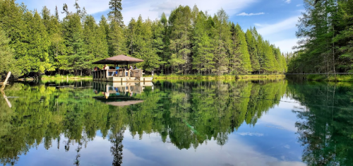 A serene lake surrounded by lush trees, with a gazebo reflecting on the calm water under a blue sky.