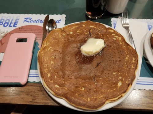 A large pancake topped with a pat of butter and syrup, served on a plate with a spoon and a pink phone nearby.