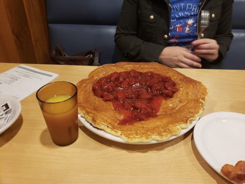 A large pancake topped with strawberry sauce sits on a table next to a glass of orange juice.