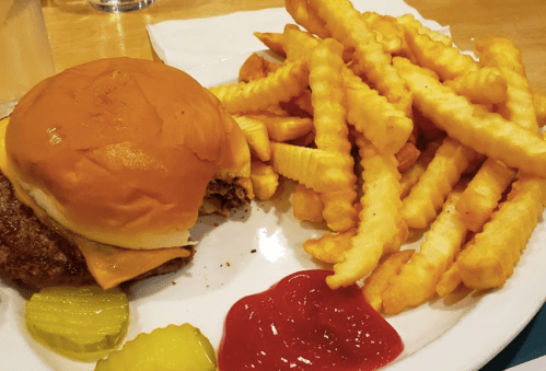 A plate with a cheeseburger, crinkle-cut fries, pickles, and ketchup. One bite taken from the burger.
