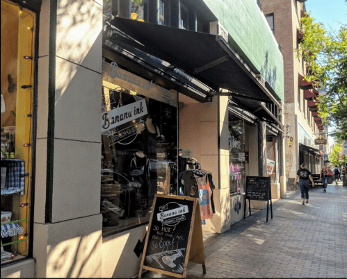 A street view of a shop called "Banana Ink" with outdoor signage and pedestrians walking by.