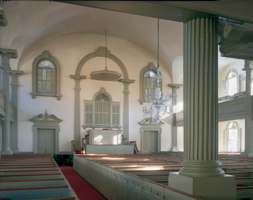 Interior of a historic church featuring wooden pews, tall columns, and a chandelier, with light streaming through windows.