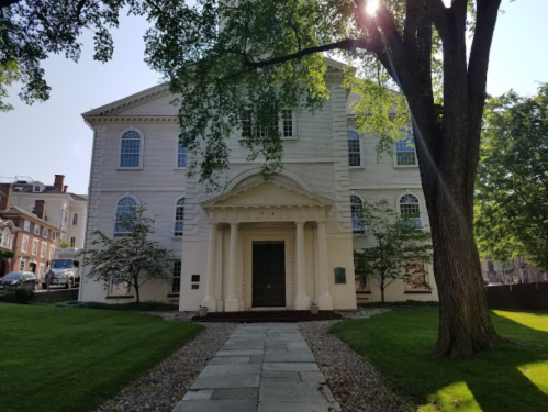 A historic white building with large windows and a portico, surrounded by green grass and trees on a sunny day.