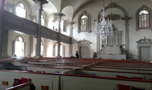 Interior of a historic church featuring wooden pews, tall windows, and a chandelier, with a person standing in the background.