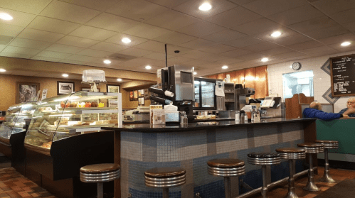 A cozy diner interior featuring a counter with stools, a display case of desserts, and patrons seated in the background.