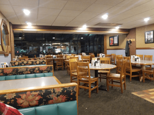 Interior of a diner with wooden tables and chairs, floral-patterned booths, and large windows showing a nighttime view.