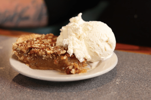 A slice of pecan pie topped with a generous scoop of vanilla ice cream on a white plate.