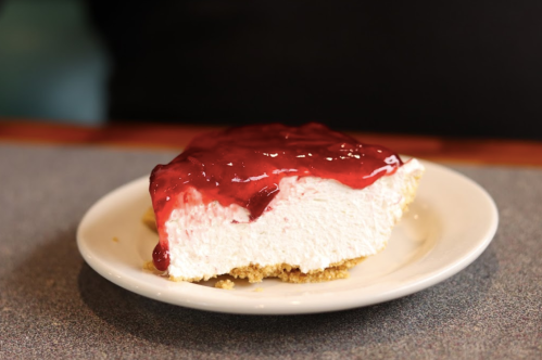 A slice of cheesecake with a glossy red fruit topping on a white plate, resting on a gray table.