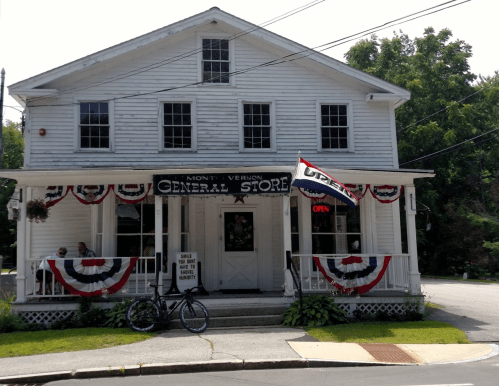 A white general store with a porch, patriotic decorations, and an "Open" sign, surrounded by greenery.