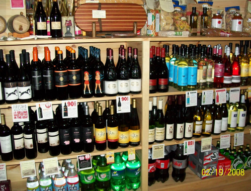 A wooden shelf displaying a variety of wine bottles and soft drinks, organized with price tags.