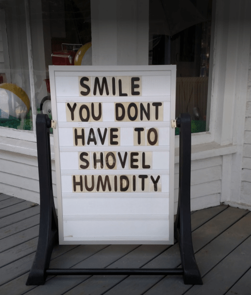 A sign reads, "SMILE YOU DONT HAVE TO SHOVEL HUMIDITY," displayed outside a building.