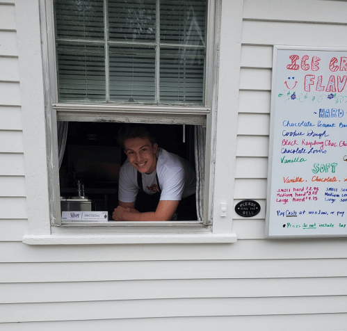 A young man smiles from a service window, with an ice cream menu displayed beside him.