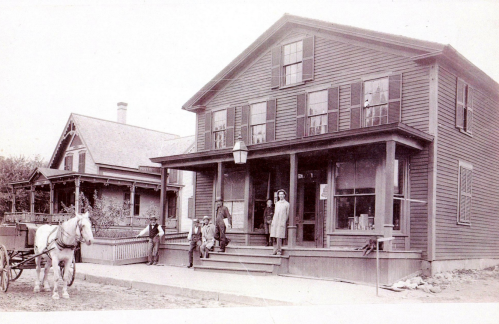 A historic black-and-white photo of a two-story house with people and a horse-drawn cart in front.