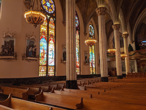 Interior of a church featuring tall stained glass windows, wooden pews, and ornate chandeliers.