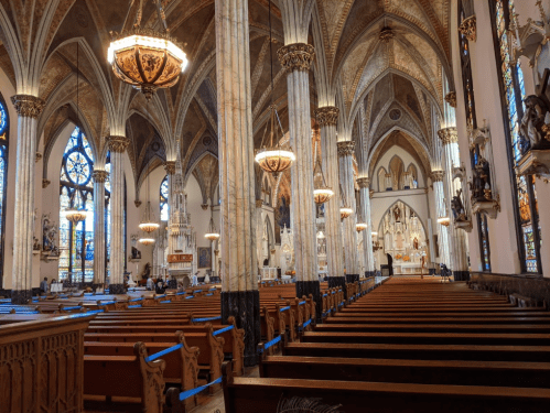 Interior of a grand church featuring tall columns, stained glass windows, and wooden pews. Soft lighting from chandeliers.