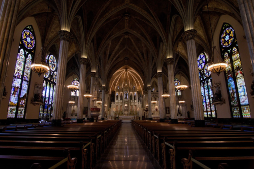 Interior of a grand church featuring tall arches, stained glass windows, and wooden pews, illuminated by chandeliers.