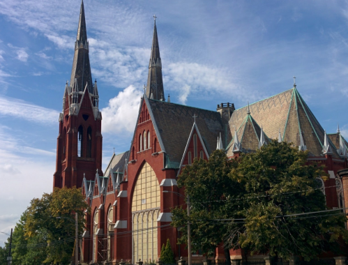 A large red brick church with tall spires and intricate architectural details under a blue sky with clouds.